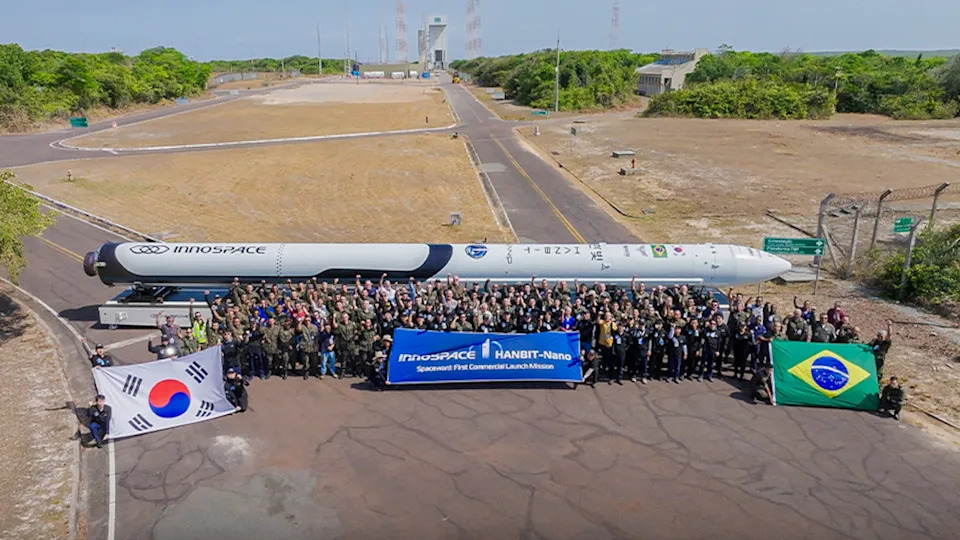 يقف نحو مائة شخص حول صاروخ أبيض منصوب أفقيًا على الأرض