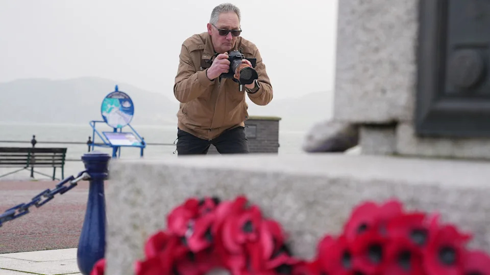 رجل ذو شعر أبيض يرتدي سترة بنية ويرتدي نظارات داكنة يميل إلى الأمام مع كاميرا ويلتقط صورة لتابوت رمادي مع إكليل من زهور الخشخاش في المقدمة