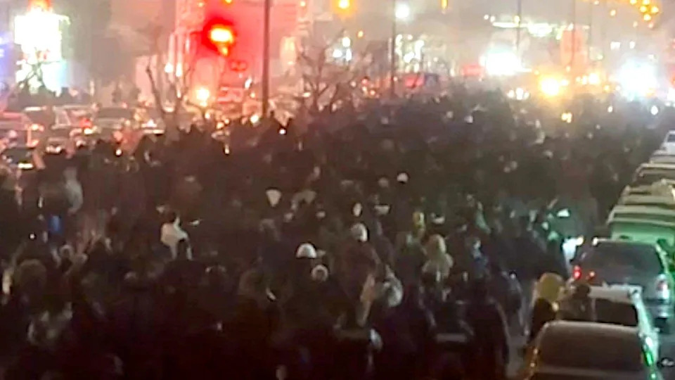 أشخاص من الخلف يسيرون على طول أحد شوارع طهران ليلاً، في 8 يناير/كانون الثاني 2026. يوجد صف من السيارات على يمين المتظاهرين، وعدة ممرات للسيارات على اليسار، مع أضواء ساطعة في المسافة.