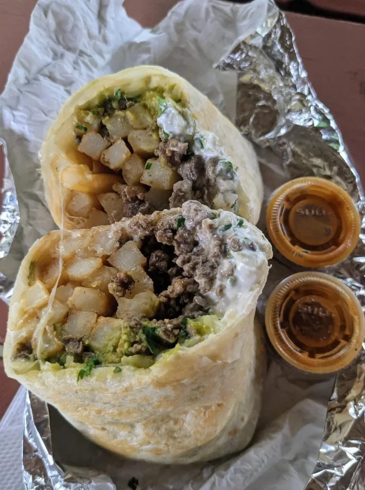 طبق يحتوي على قطعتين من البوريتو، نصف حشوة من الأرز والفاصوليا والخضروات، تقدم مع صلصات غموس على سطح مغطى بورق الألمنيوم. رديت عرض آخر