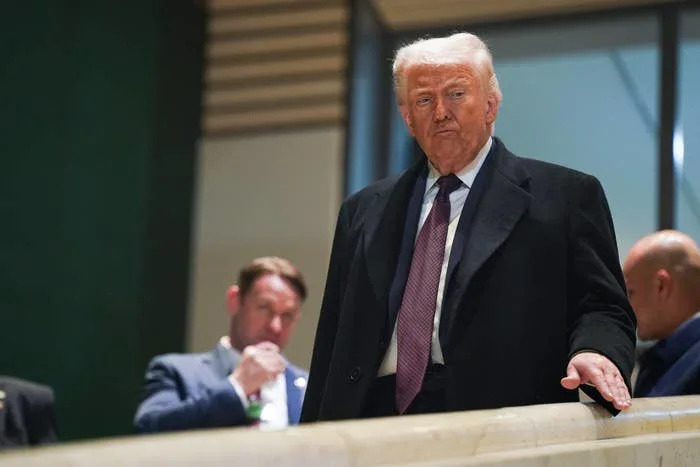 رجل يرتدي بدلة ومعطفًا ينظر إلى الأسفل من الشرفة، ويحيط به آخرون يرتدون ملابس العمل