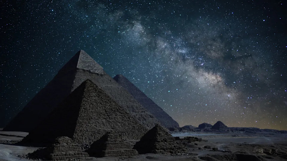 سلسلة من الأهرامات البنية على خلفية صخرية مع سماء ليلية زرقاء مرصعة بالنجوم فوقها.