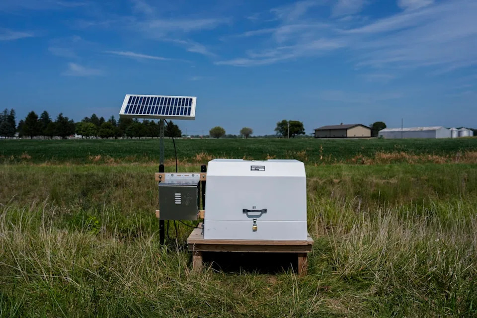 يوجد صندوق أبيض صغير في حقل من العشب، وخلفه لوح شمسي.