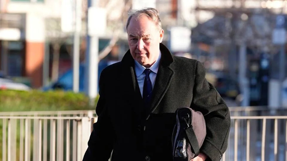 رجل ذو شعر بني أبيض يمشي مرتديًا بدلة ومعطفًا طويلًا داكنًا 