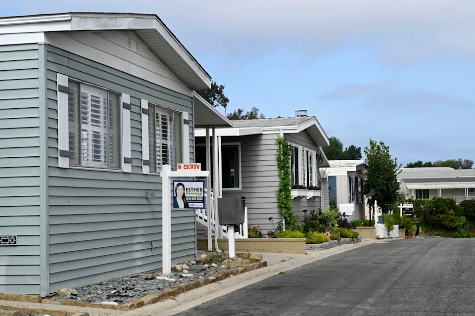 تصطف المنازل المتنقلة على طول شارع هادئ، ويعرض أحدها لافتة عقارية للبيع، وتظهر الأشجار والشجيرات حول المنطقة