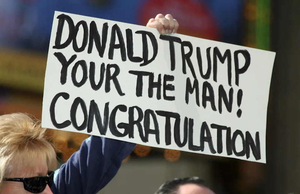 شخص يحمل لافتة تقول "دونالد ترامب أنت الرجل! تهانينا" في مناسبة عامة، محاطًا بأشخاص آخرين