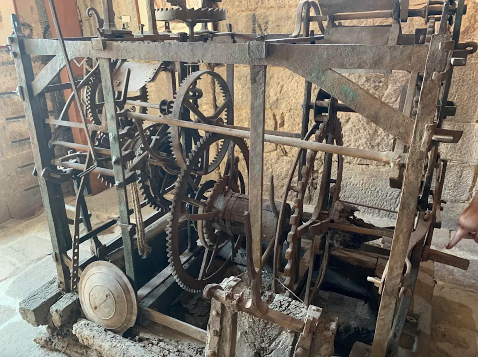 آلية الساعة العتيقة مع التروس والأوزان، معروضة في محيط بجدران حجرية