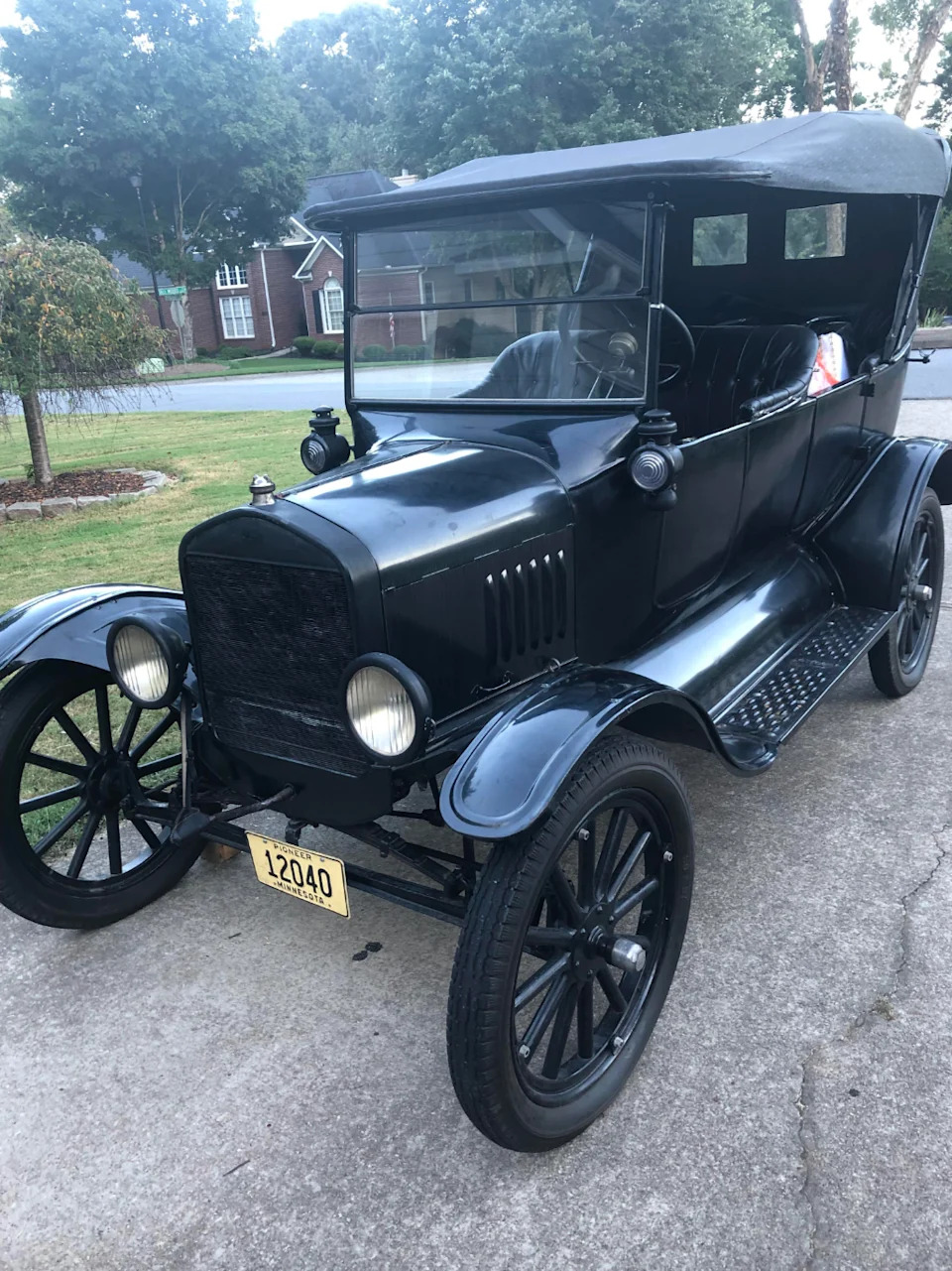 سيارة قديمة متوقفة في الشارع، تتميز بتصميم كلاسيكي بعجلات كبيرة وسقف مظلي. تظهر الأشجار والمنازل في الخلفية