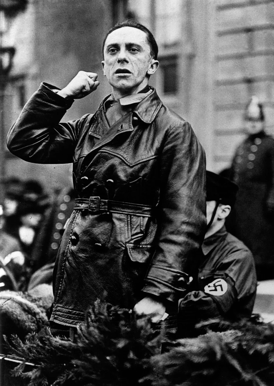 شخصية تاريخية ترتدي معطفًا جلديًا ترفع قبضتها أثناء التحدث إلى حشد من الناس، مع وجود شارة مرئية على ذراع شخص واحد قريب