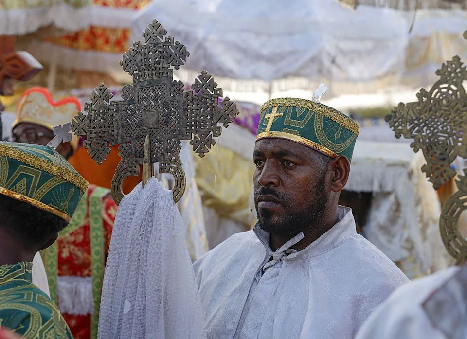 يقف كاهن بالملابس التقليدية ويحمل صليبًا.