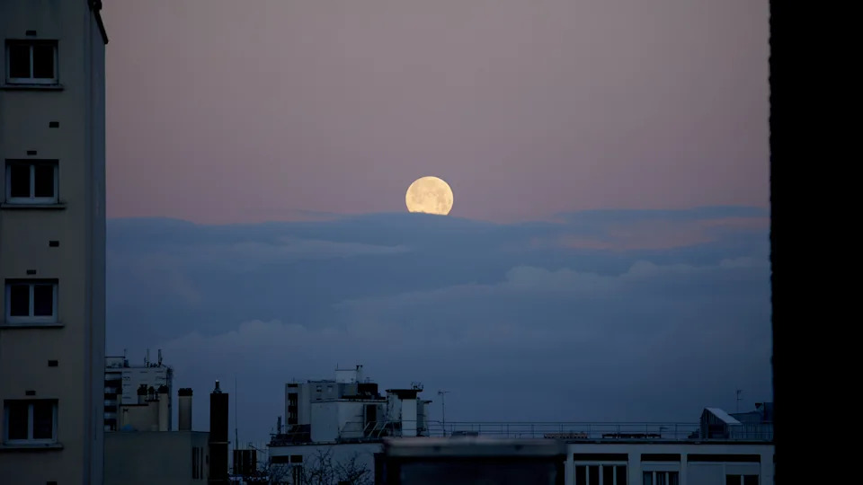 تم تصوير البدر وهو يرتفع فوق سحابة كثيفة في سماء المساء الزرقاء الداكنة بين المباني.