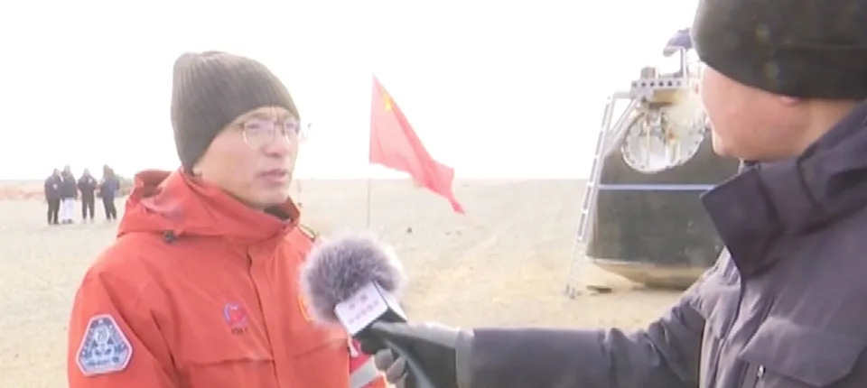 رجل يرتدي سترة حمراء وقبعة سوداء مع نظارات يجيب على شخص يرتدي سترة سوداء ويحمل ميكروفونًا