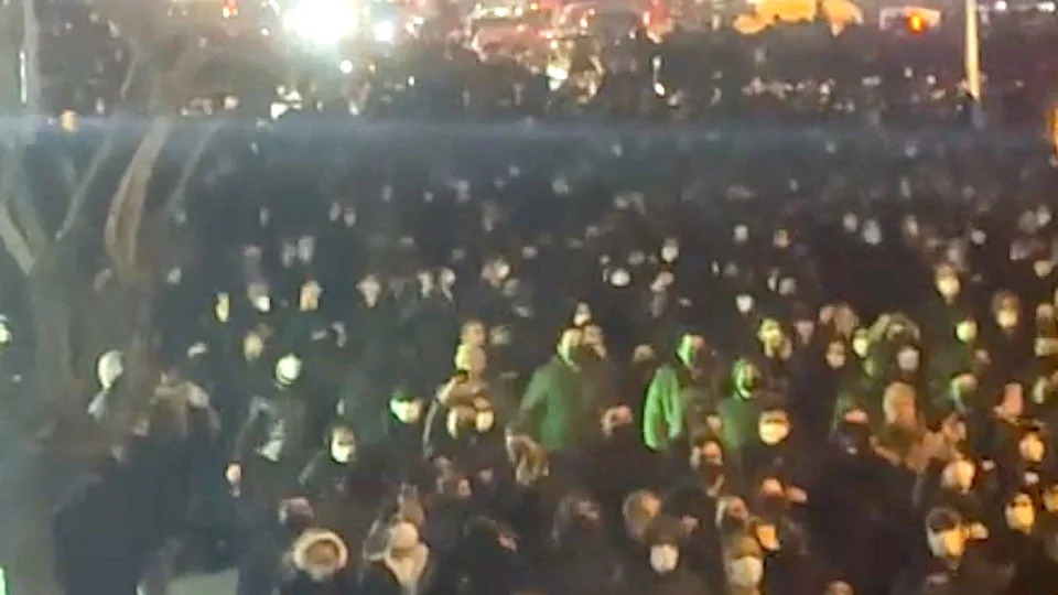 تجمعت مجموعة كبيرة من الناس في أحد الشوارع ليلاً. يرتدي معظمهم أقنعة والعديد من القلنسوات. يتدفق الضوء إلى الداخل، ويضيء شجرة، مع أضواء ساطعة في الخلفية.