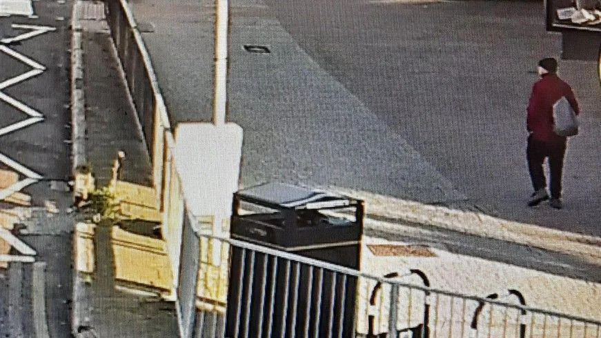 تظهر صورة CCTV رجلاً يسير على رصيف واسع بجوار الطريق. يرتدي سترة باللون العنابي، وقبعة داكنة، وسروالًا وحذاء داكنًا، ويحمل حقيبة على كتف واحد. 