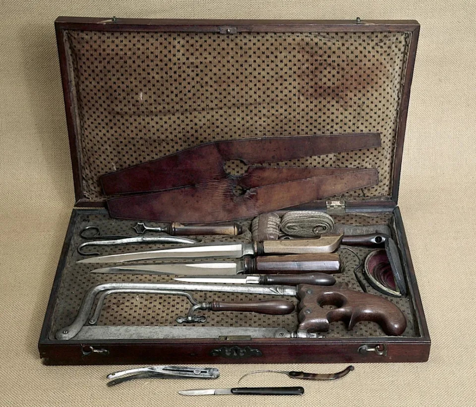 مجموعة جراحية كلاسيكية مفتوحة تحتوي على أدوات طبية متنوعة، بما في ذلك المناشير والمشارط والمشابك، معروضة في علبة خشبية مبطنة