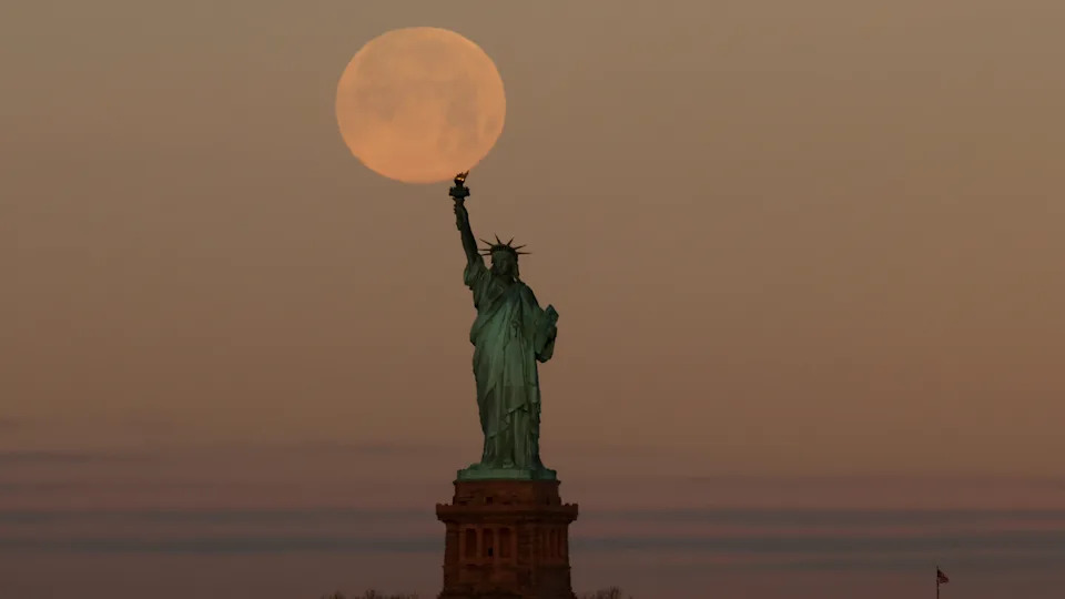 تم تصوير البدر باللون الأصفر البرتقالي وهو يلمس شعلة تمثال الحرية في نيويورك في سماء مسائية ضبابية. 
