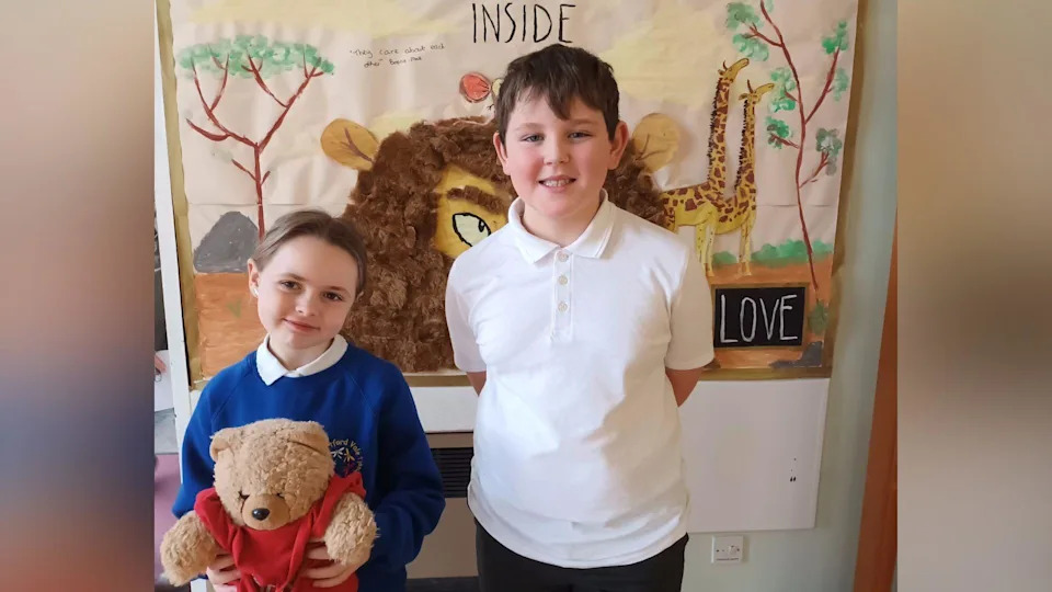 اثنان من تلاميذ المدارس الابتدائية يقفان أمام لوحة العرض المدرسية. بودي يحمل دمية دب، وأوليفر يقف ويداه خلف ظهره. 