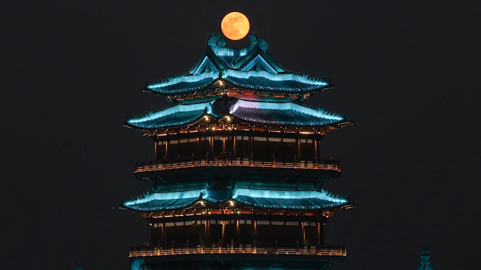 تم تصوير البدر متوهجًا فوق سطح مبنى صيني تقليدي في سماء الليل المظلمة. تمت إضاءة سطح المبنى لإبراز معالمه المزخرفة.