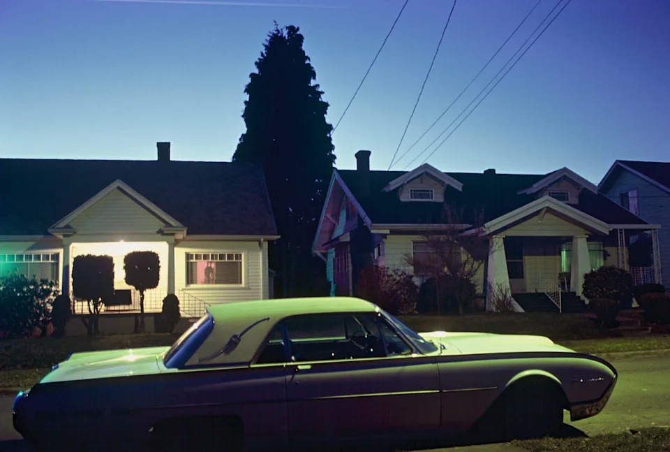 تقف سيارة عتيقة في أحد شوارع الضواحي عند الغسق، مع منزلين مريحين مضاءين بهدوء في الخلفية