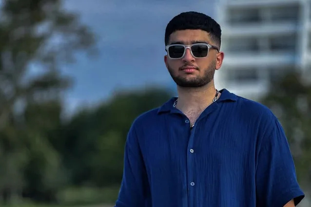 شاب يقف في الهواء الطلق بالقرب من حاجز معدني، ويرتدي نظارة شمسية داكنة بإطارات فاتحة اللون، وقميصًا أنيقًا أزرق داكن بأكمام قصيرة وأزرار، وجينز أسود، وساعة على معصمه الأيسر، وقلادة على شكل سلسلة. وله لحية سوداء قصيرة تمتد من أذنيه إلى ذقنه. وخلفه يوجد حقل عشبي به أشجار خضراء مورقة ومبنى طويل أبيض حديث متعدد الطوابق يمكن رؤيته جزئيًا. السماء ملبدة بالغيوم، مع مزيج من السحب الزرقاء والرمادية.