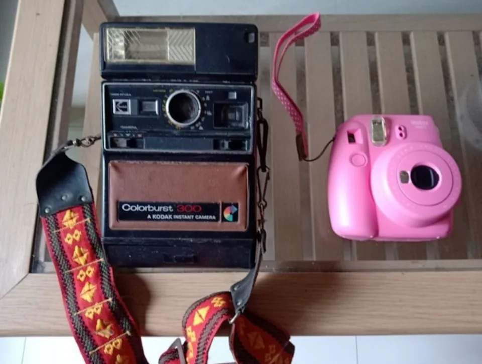 كاميرتان فوريتان على طاولة: كاميرا كوداك قديمة بحزام وكاميرا Instax وردية اللون حديثة