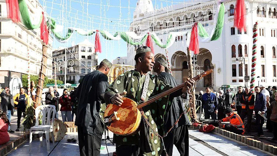 رجل يرتدي سترة خضراء يعزف على آلة تشبه الجيتار أمام جمهور في الهواء الطلق
