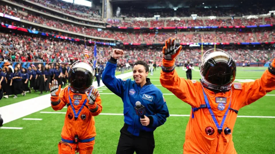 امرأة ذات شعر داكن قصير ترتدي بدلة زرقاء وتحمل ميكروفونًا ويحيط بها شخصان يرتديان بدلات فضائية برتقالية وخوذات داكنة في ملعب كرة قدم مع ساحة كاملة خلفهما.