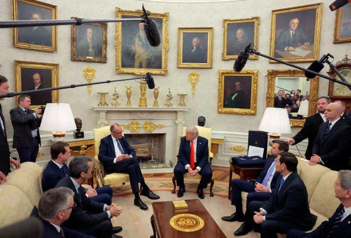 اجتماع في مكتب رسمي مع أشخاص جالسين ومحاطين بلوحات شخصية وميكروفونات في الأعلى