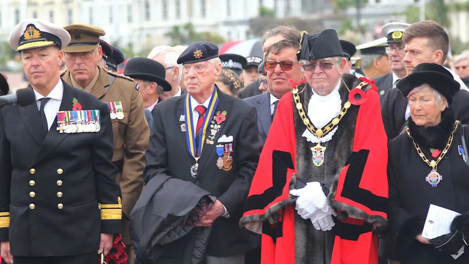 رجل يرتدي زي أميرال يقف رسميًا. يرتدي قبعة بيضاء وسترة بحرية وقميصًا أبيض وربطة عنق. يوجد على صدره سلسلة من الميداليات معلقة بأشرطة ملونة، وعلى جانبه يتدلى مقبض السيف الذهبي اللامع. 