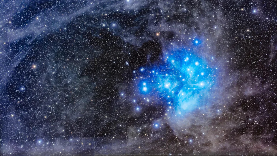 تم تصوير مجموعة من النجوم الزرقاء والبيضاء محاطة بتوهج أزرق منتشر مقابل حقل النجوم الخافت خلفها، والذي تتخلله سحب ناعمة.