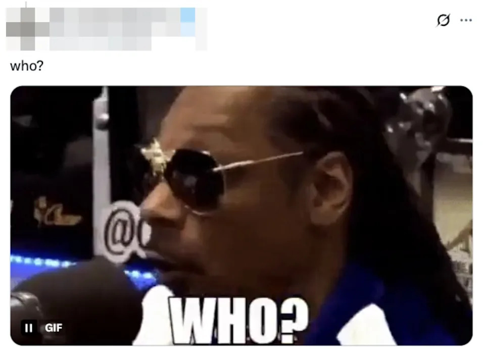 شخص ذو شعر طويل ونظارة شمسية يتحدث في الميكروفون. يقرأ النص، "من؟"