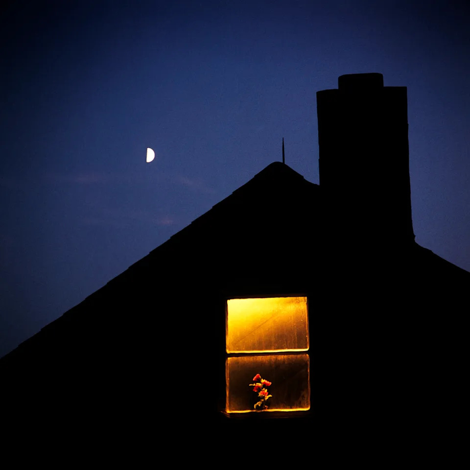 نافذة ذات إضاءة خافتة بها زهور تقع تحت هلال مقابل سماء مظلمة، فوق سقف مظلل