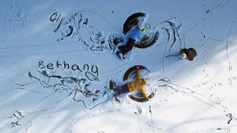 الأطفال يصنعون ملائكة الثلج في الثلج - يحركون أذرعهم وأرجلهم ذهابًا وإيابًا أثناء الاستلقاء على الجليد ليصنعوا أشكال الأجنحة. هناك ثلاثة أطفال وجميعهم يرتدون ملابس دافئة وقبعات صوفية