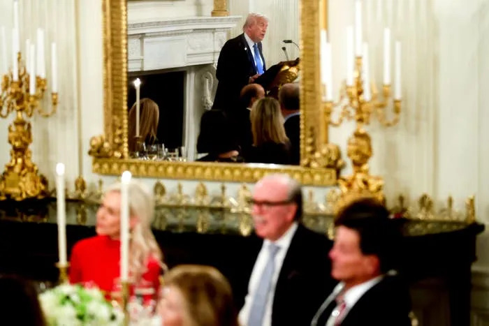 رجل يتحدث على منصة في قاعة مناسبات رسمية بها مرآة مزخرفة. يجلس الضيوف على الطاولات في المقدمة