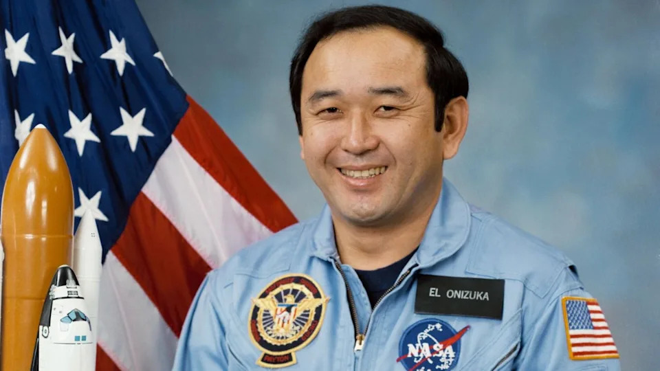 رجل يرتدي بدلة طيران زرقاء يقف لالتقاط صورة بجانب العلم الأمريكي ونموذج لصاروخ